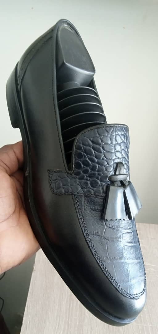 Il s'agit d'une paire de mocassins noirs à pompons (en anglais tassel loafers) pour hommes. Ces chaussures présentent un style slip-on avec deux pompons décoratifs sur la tige et un motif crocodile ou floral en relief sur le cuir.